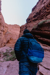 A man wearing a black jacket and a blue backpack is walking through a canyon
