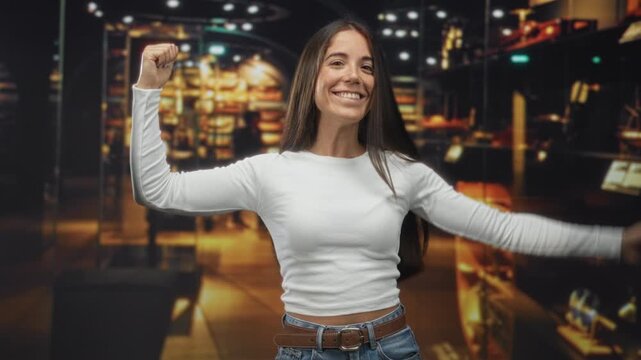 Young woman points to flexed bicep inside a building, smiling and flexing arm while wearing white top and jeans; confidence empowerment.