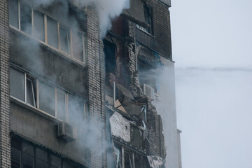 A multi-story residential building exhibiting severe structural damage, broken balconies, and thick smoke following a fire or explosion. High-pressure water is pouring onto the damaged brick façade, a © Denis Chubchenko