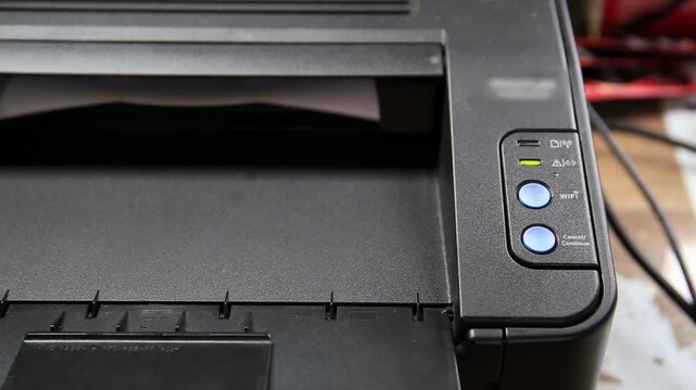 Close up view of black laser printer operating, white paper emerges from output tray with printed text. Indicator lights illuminate during print job. Essential office equipment.