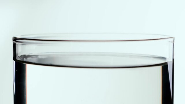 A single liquid droplet falls into clear water, creating soft concentric ripples that gradually settle in a bright minimalist glass composition on a neutral background with subtle diffusion below.