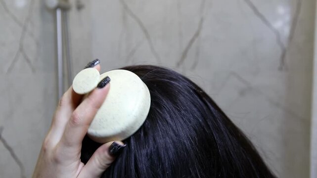 Close-up of hand massaging scalp with round brush. Long dark hair receives treatment indoors. Bathroom setting, part of daily routine for hair health, beauty.
