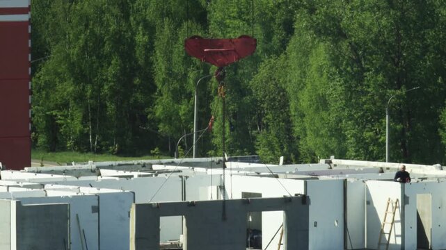Construction site activity. Precast concrete wall panels. Building structure formed. Red crane hook positions elements. Workers ensure panel placement. Green forest background.