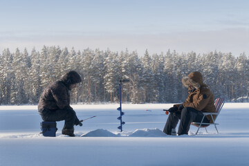 Two fishermen ice fishing on a frozen forest lake in winter. Snow, ice and cold weather create a calm natural outdoor scene. Seasonal winter activity and traditional leisure concept.