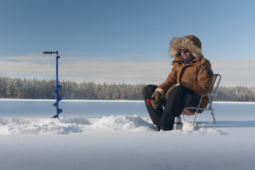 Winter fisherman sitting on ice during ice fishing. Cold outdoor recreation with auger, fishing rod and frozen lake landscape.