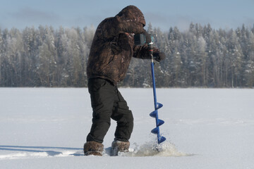 Man drilling ice hole with electric auger on a frozen lake. Winter fishing activity in cold outdoor nature, modern ice fishing equipment and seasonal lifestyle