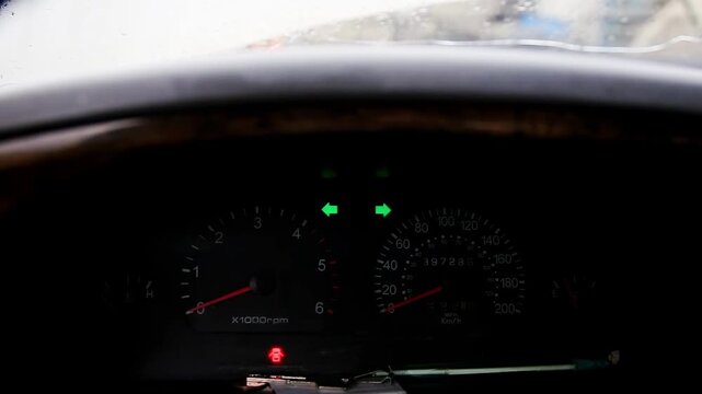 Car dashboard. Instrument cluster active. Green hazard lights blink. Red warning light glows. Speedometer, tachometer, odometer show vehicle data. Highlights alerts, operational status.