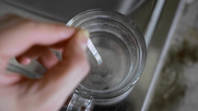 Close up view of person hand holding simple test strip, immersing into clear glass of tap water. Household method determines mineral content, alkalinity, pH level, chlorine, nitrites, nitrates, hardne