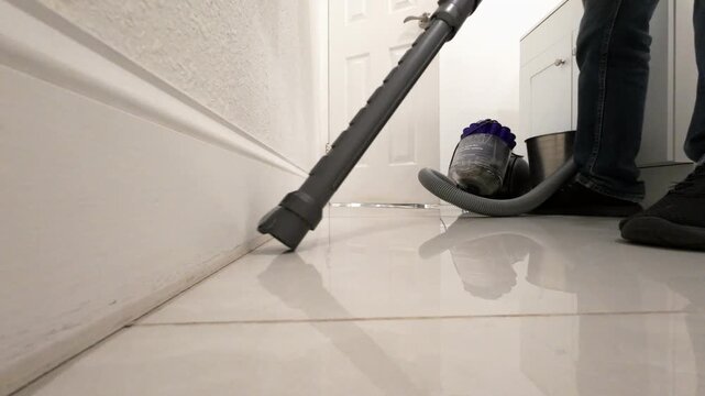 Vacuum cleaner moving back and forth along a white baseboard while cleaning the floor. Low angle floor level view showing vacuum head, black sneakers and legs in jeans during home cleaning.
