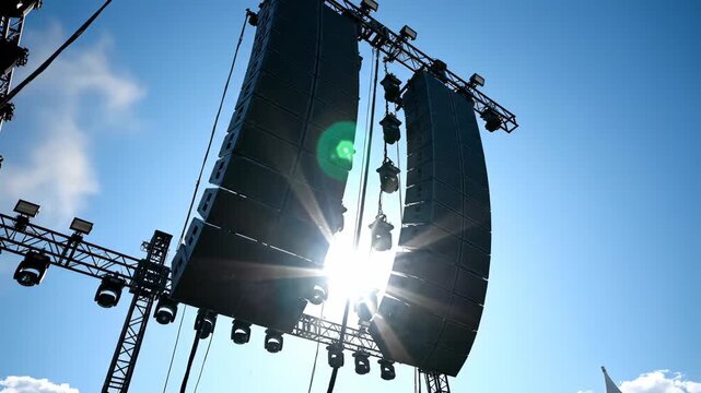 The sun radiates brilliantly through massive sound speakers at an outdoor music festival, casting a dazzling glare against a vibrant blue sky filled with wispy clouds. The scene is alive with energy.