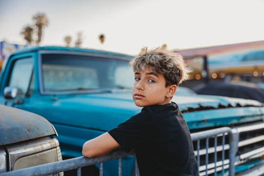 Boy leaning on metal fence with vintage cars.