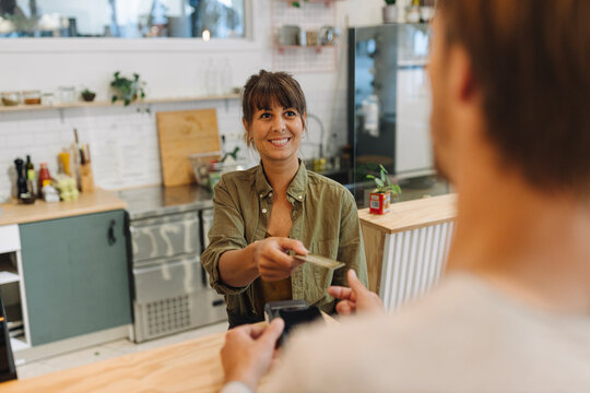 Smiling female owner giving back credit card to male customer at checkout in coffee shop