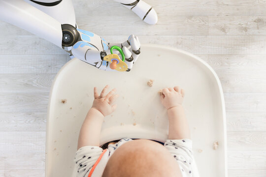 Robot hand giving pacifier to baby boy sitting in high chair playing with bread crumps, top view