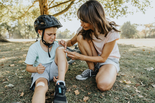 Mother putting bandage on son's knee while sitting in public park during sunny day