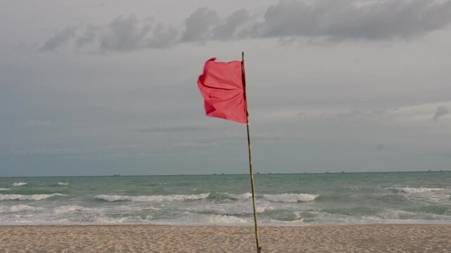 Red warning flag flutters rapidly in strong seaside wind above empty beach with choppy waves and cloudy sky creating dramatic safety signal  in coastal environment