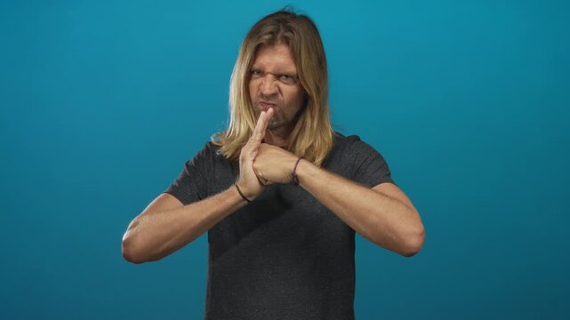 Blond man with long hair grimaces and presses palms together against a blue studio backdrop; determination grit.