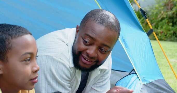 After pitching blue tent, African American father and son sharing stories to bond on yard grass