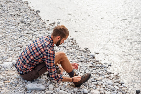Man tying boot on rocky riverbank during summer hiking adventure