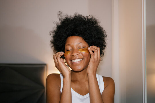 Smiling woman in pyjamas applying eye patches in the bedroom
