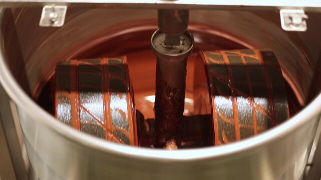 Close up rotating industrial cocoa beans mixing rollers stirring and coating with glossy melted chocolate inside factory for confectionery production.