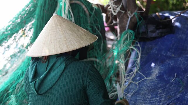 Back view of viet woman in conical hat slowly pulling and untangling green fishing nets on a blue tarp at vietnamese village, hands working in sequential motion for coastal livelihood.