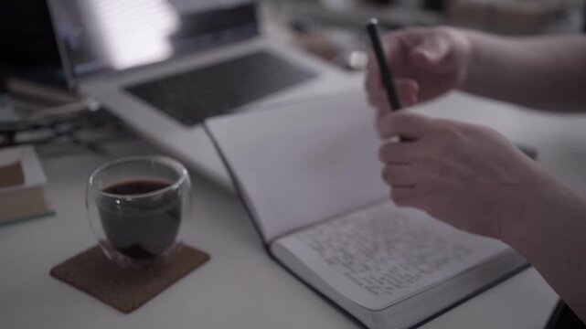 Close-up of person writing in notebook with coffee during evening at desk. Concept of productivity, journaling, calm routine and focused work.