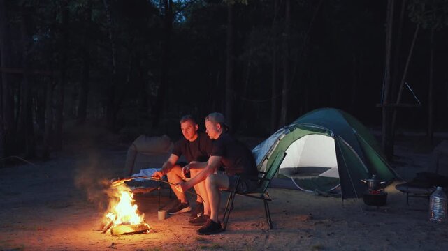 Serene evening around campfire. Calm nighttime setting with two men enjoying peaceful stargazing. Quiet outdoor environment with individuals relaxing by gentle campfire beneath pine trees