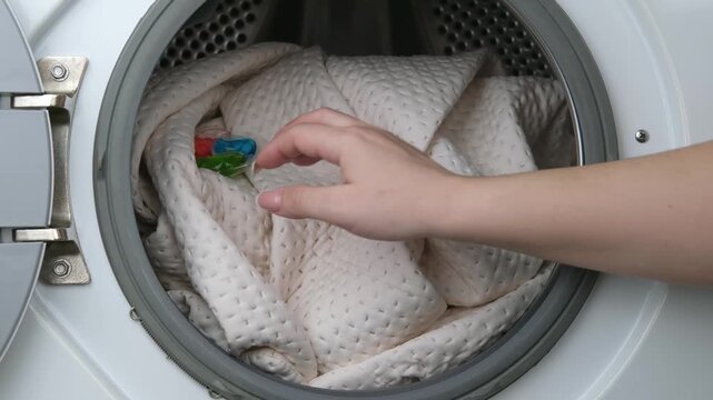 A woman's hand places a powder capsule on a beige blanket in a washing machine
