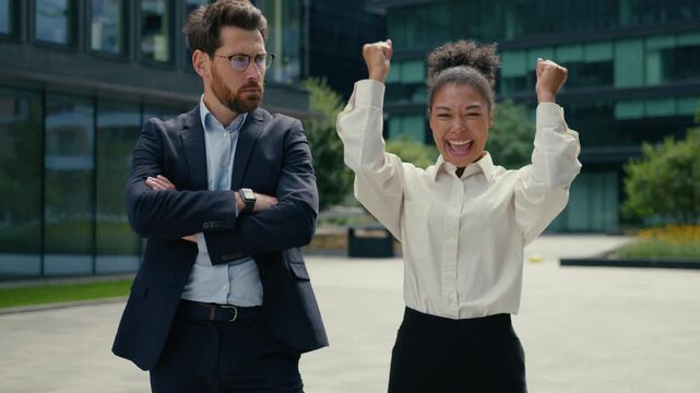 Happy successful African businesswoman celebrating win in city Caucasian businessman jealous different emotions colleagues joy success envy offended male female diversity workplace tension outdoors