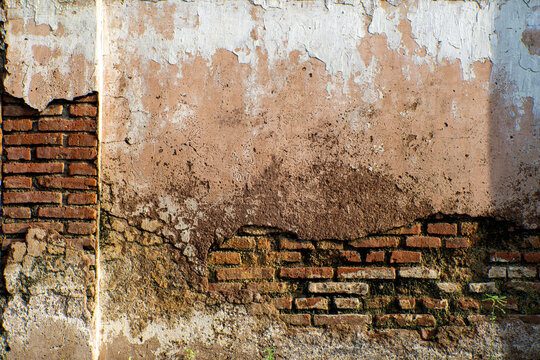 Weathered red brick wall texture with peeling plaster and moisture stains. Grungy vintage brickwork background for industrial and architectural design in rural Indonesia.