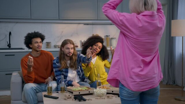 Multiracial friends playing charades guessing game at home room Caucasian woman gesturing explaining word fun entertainment diverse African women and men pantomime joyful group laugh interact together