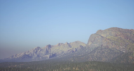 A breathtaking view of a mountain range features rugged peaks and a vibrant blue sky. Lush evergreen trees fill the valley below, creating a serene natural landscape.