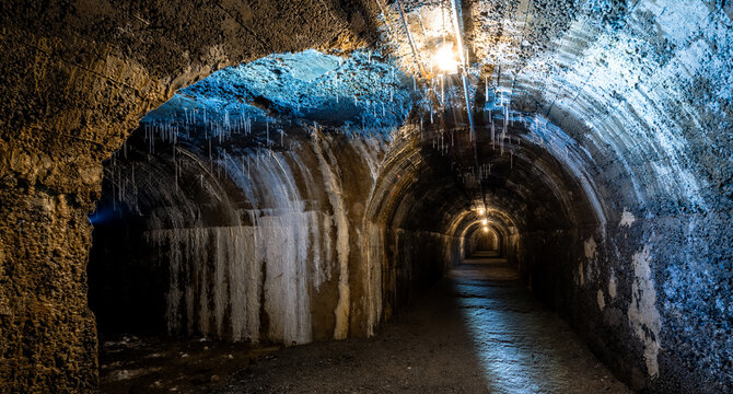 Corridors in a historic bunker at an underground junction. A lost place with sparse incandescent lighting, damp walls, and stalactites forming on the concrete vaulted ceilings.  Eerie atmosphere.