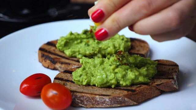 Woman's hand squeezing a lemon wedge over grilled rye bread toast with creamy guacamole and fresh cherry tomatoes, preparing a healthy and delicious vegetarian breakfast or snack at home
