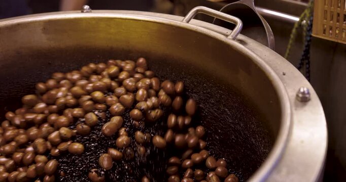 Roasted chestnuts tumbling inside large pan over fire at street market, vendor stirring nuts with rhythmic motion, with glossy reflecting orange flame in slowmotion, Thai street snack making
