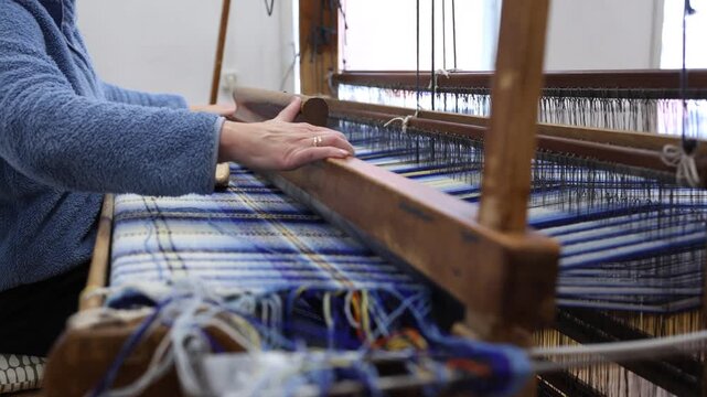 Close-up of a hand guiding blue and white threads on a traditional wooden loom, creating a plaid textile pattern in a craft workshop