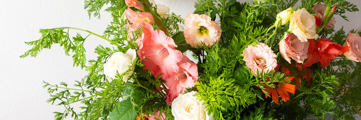A bouquet of roses, gladioli, zinnias, and eustomas stands in water. Carrots with green tops are part of the arrangement on a plain table against a light background, banner © pundapanda