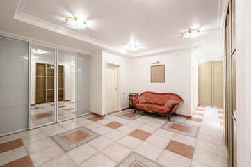 Fototapeta premium Spacious hallway with tiled floor, red patterned sofa, mirrored wardrobe, recessed ceiling lights, framed blank picture, light-colored walls, wooden doors