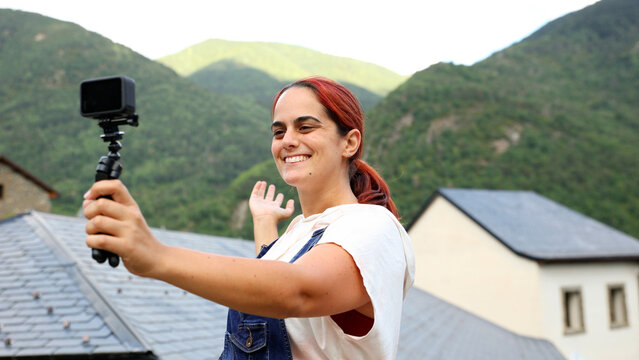 Young woman filming travel vlog with action camera on tripod in mountain village, sharing adventure content outdoors