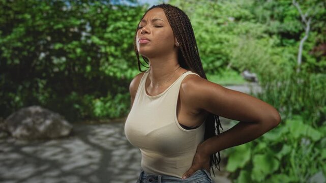 Woman holding lower back for backache on a forest path, eyes closed and leaning back in pain; pain resilience.