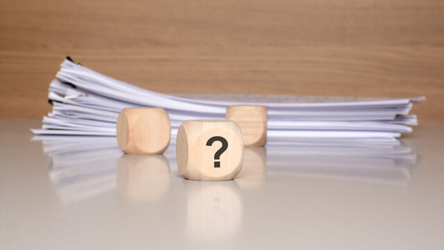 Question mark on wooden cube with stack of documents. Business uncertainty, legal questions and paperwork analysis concept.