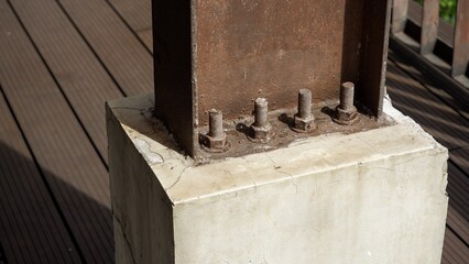 A close-up of rusted steel I-beam anchored to a concrete foundation with heavy-duty bolts and nuts. A detailed view of industrial structural engineering on a wooden deck background. © agung