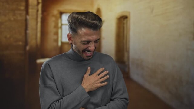 Young hispanic man with hand on chest while laughing amid worn stone walls in an old building; pure joy.