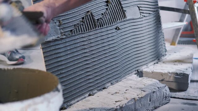 Master tiler spreads glue on huge large format tiles of XXL size. Notched trowel, professional tile work, bathroom renovation, laying blue tiles, slow motion