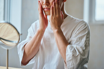 Asian young man applying skincare in front of mirror (face cropped)