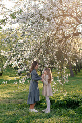 Fototapeta premium A woman and a girl stand under a blooming cherry tree. The woman has long brown hair and wears a gray dress. The girl has blonde hair and wears a light dress.
