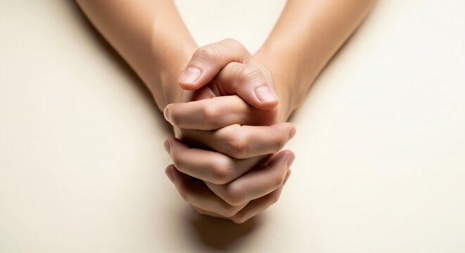 Clasped hands in a prayer or pleading gesture, fingers tightly interlocked, plain cream background.