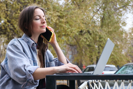 Mid adult woman working remotely on laptop while talking on smartphone at outdoor cafe terrace in autumn park