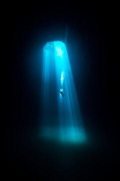 View of a diver descending through a sunlit cenote, the turquoise rays cutting through the dark water, creating an ethereal underwater scene, Quintana Roo, Mexico.