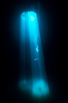 View of sunbeams piercing the dark depths as divers descend on a rope into the cool, blue waters of a cenote, Quintana Roo, Mexico.